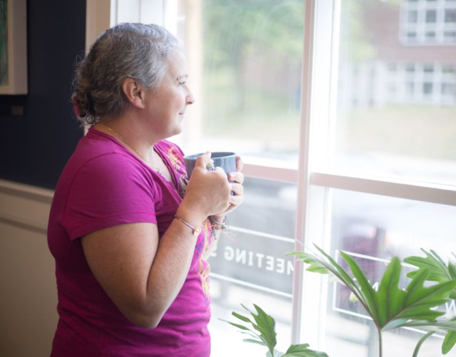 A woman stands by a window with a cup, looking outside